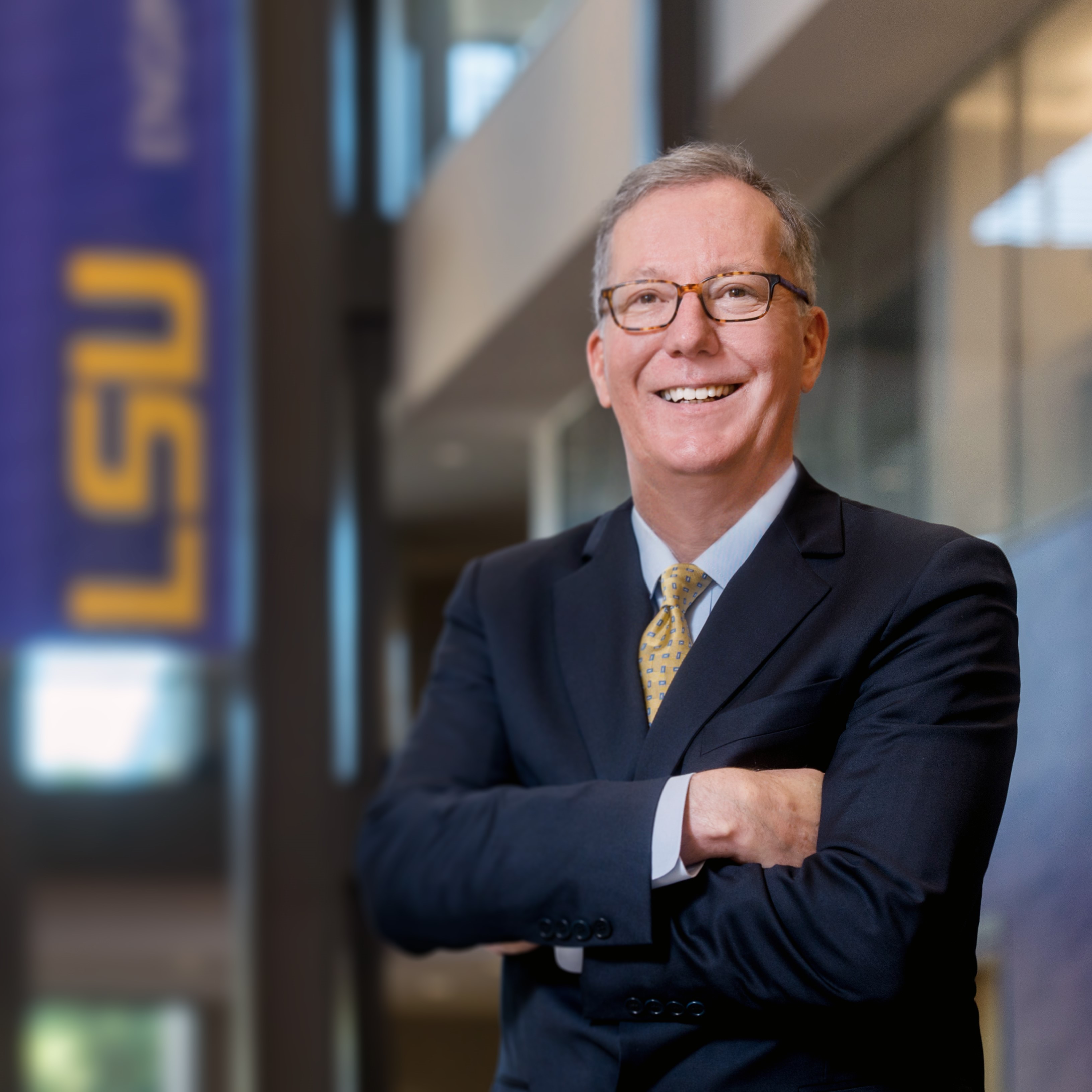 Brad Ives has his arms crosse over his chest. He is wearing a dark suit and there is an LSU flag in the background. 